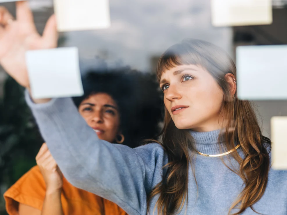 Girl in blue turtle neck sweater looks at post-it notes on glass board with fellow colleague. 