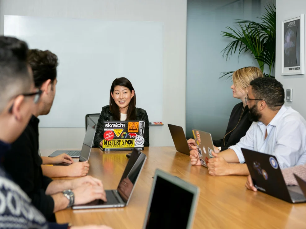 Young Asian woman sits in front of laptop covered in cool stickers, and talks to colleagues sat around wooden table in modern office environment.